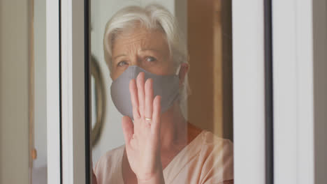 Senior-caucasian-woman-wearing-face-mask-looking-out-of-window-at-home