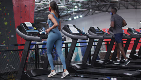 Fit-caucasian-woman-and-african-american-man-wearing-face-masks-running-on-treadmill-in-the-gym
