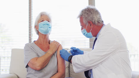 Male-caucasian-doctor-wearing-face-mask-giving-injection-to-senior-caucasian-woman-at-hospital