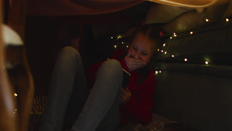 Caucasian-girl-smiling-while-using-smartphone-under-blanket-fort-during-christmas-at-home