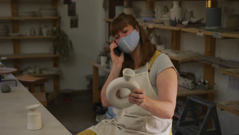 Female-caucasian-potter-wearing-face-mask-and-apron-holding-pot-and-talking-on-smartphone-at-pottery
