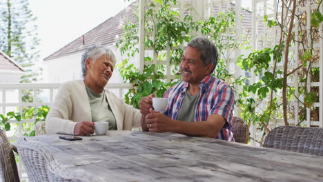 Happy-senior-mixed-race-couple-having-coffee-talking-in-garden