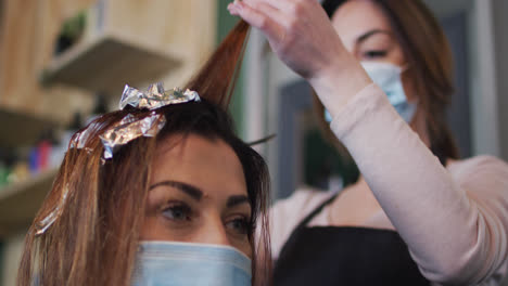 Female-hairdresser-dying-hair-of-female-customer-wearing-face-mask-at-hair-salon
