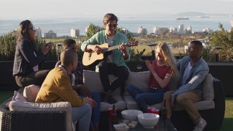 Young-Caucasian-man-playing-guitar-on-a-rooftop-with-his-friends