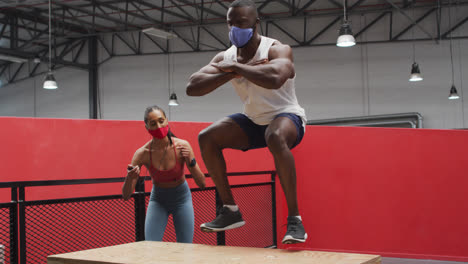 African-american-man-jumping-on-box-wearing-face-mask-at-gym,-mixed-race-woman-timing