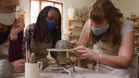 Diverse-potters-wearing-face-masks-and-aprons-working-on-pot-on-potters-wheel-at-pottery-studio