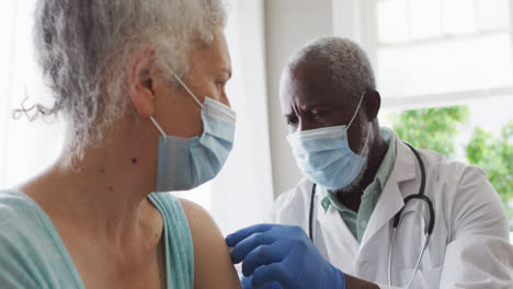 Male-african-american-doctor-wearing-face-mask-injecting-covid-19-vaccine-into-senior-female-patient