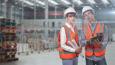 Two-Caucasian-workers-in-a-warehouse-with-oranges-high-vest-and-hat-area-talking-and-checking-notes