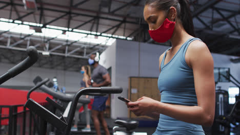 Mixed-race-woman-wearing-face-mask-using-smartphone-at-gym