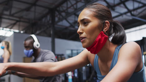 Mixed-race-woman-wearing-lowered-face-mask-exercising-at-gym