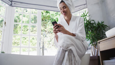 Mixed-race-woman-sitting-on-bathtub-and-using-her-smartphone-in-bathroom