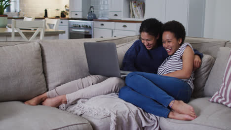 Mixed-race-lesbian-couple-sitting-on-couch-embracing-and-using-laptop