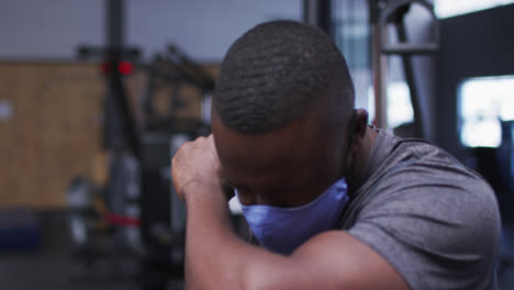 Fit-african-american-man-wearing-face-mask-sneezing-on-his-elbow-in-the-gym