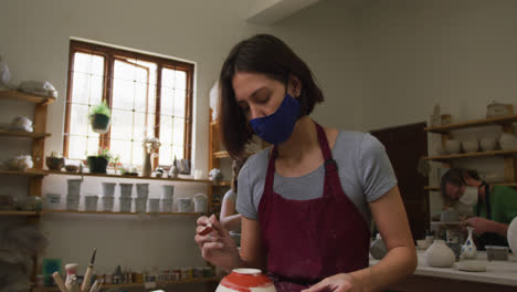Female-caucasian-potter-wearing-face-mask-and-apron-using-glaze-brush-to-paint-pot-on-potters-wheel-