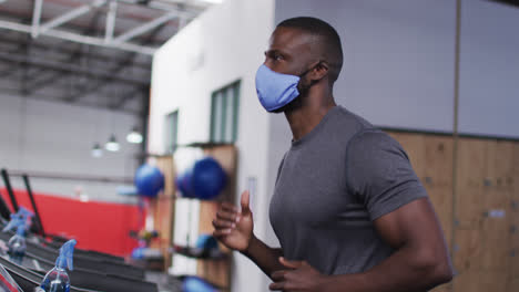 Fit-african-american-man-wearing-face-mask-running-on-treadmill-in-the-gym