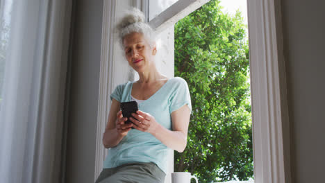 Ältere-Kaukasische-Frau,-Die-Zu-Hause-Ein-Smartphone-Am-Fenster-Benutzt