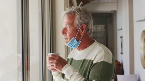 Senior-caucasian-man-with-lowered-face-mask-drinking-coffee-while-looking-out-of-window-at-home