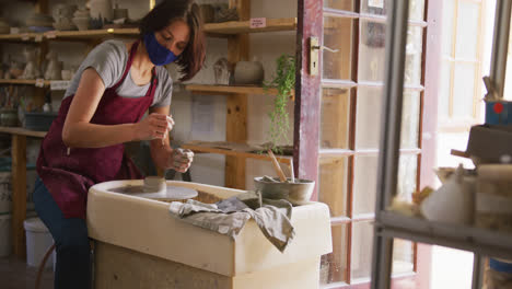 Female-potter-wearing-face-mask-and-apron-creating-pottery-on-potters-wheel-at-pottery-studio