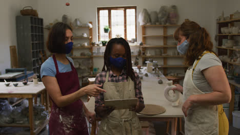 Diverse-female-potters-wearing-face-mask-and-apron-using-digital-tablet-at-pottery-studio