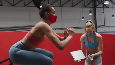 Mixed-race-woman-jumping-on-box-wearing-face-mask-at-gym,-caucasian-woman-timing