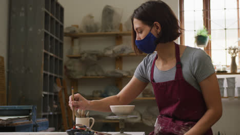 Female-potter-wearing-face-mask-and-apron-painting-pot-on-potters-wheel-at-pottery-studio
