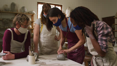 Diverse-potters-wearing-face-masks-and-aprons-working-on-clay-at-pottery-studio