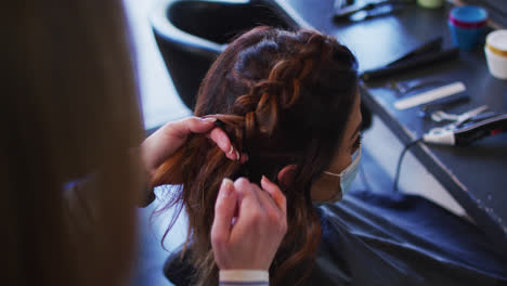 Female-hairdresser-braiding-hair-of-female-customer-wearing-face-mask-at-hair-salon