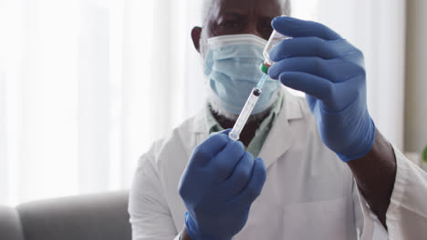 Male-african-american-doctor-wearing-face-mask-pulling-out-covid-19-vaccine-into-syringe
