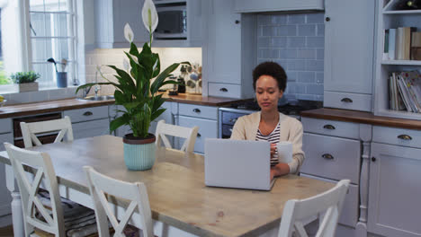 Mixed-race-woman-using-laptop-and-drinking-coffee-in-kitchen