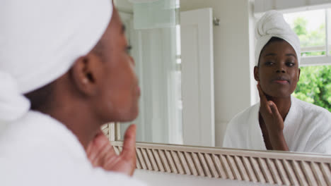 Afroamerikanische-Frau-Im-Bademantel-Trägt-Gesichtscreme-Auf,-Während-Sie-Zu-Hause-In-Den-Spiegel-Schaut