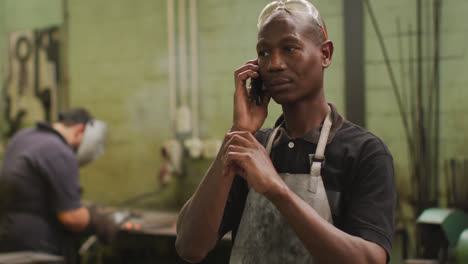 African-American-male-factory-worker-at-a-factory-standing-in-a-workshop,-talking-on-a-smartphone