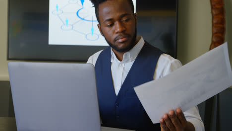 Front-view-of-young-black-businessman-looking-at-documents-while-using-laptop-in-a-modern-office-4k