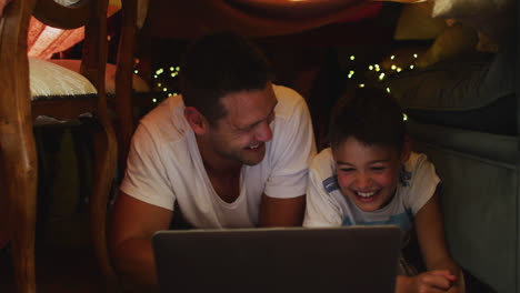 Caucasian-father-and-son-smiling-while-using-laptop-under-blanket-fort-during-christmas-at-home