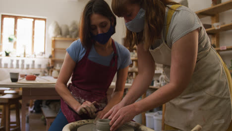 Two-female-caucasian-potters-wearing-face-masks-and-aprons-creating-pottery-on-potters-wheel-at-pott