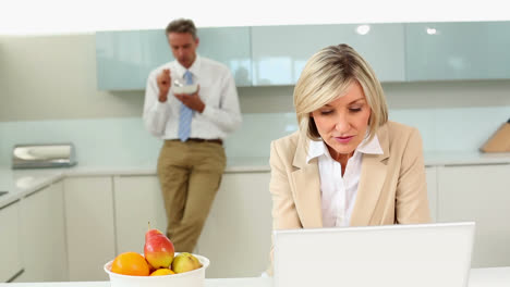 Businesswoman-using-laptop-while-husband-eats-cereal-before-work