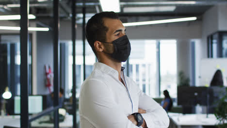 Portrait-of-mixed-race-businessman-wearing-face-mask-standing-in-office-looking-to-camera