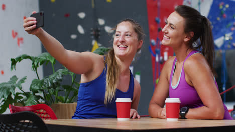Two-happy-caucasian-women-taking-selfie-with-smartphone-in-cafe-at-indoor-climbing-wall