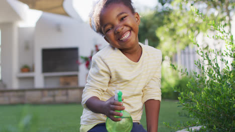 Porträt-Eines-Glücklichen-Afroamerikanischen-Mädchens,-Das-Im-Garten-Pflanzen-Gießt-Und-Eine-Sprühflasche-Hält