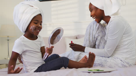 Happy-african-american-mother-and-daughter-wearing-towels-sitting-on-bed-looking-at-mirror