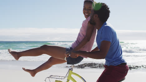 African-american-couple-riding-on-bike-on-the-beach,-woman-sitting-on-handlebars