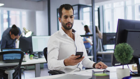 Mixed-race-businessman-sitting-at-desk-using-computer-and-smartphone-in-busy-office