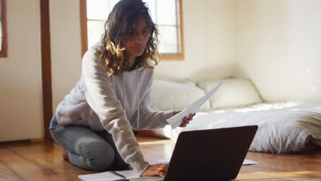 Mixed-race-woman-working-at-home,-kneeling-on-floor-using-laptop-and-holding-paperwork-in-cottage