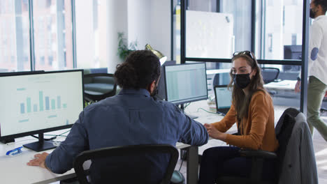 Diverse-male-and-female-colleague-at-desks-using-computers-wearing-face-masks-and-elbow-bumping