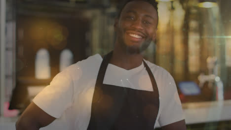 Spots-of-light-against-portrait-of-male-barista-wearing-apron-smiling-at-cafe