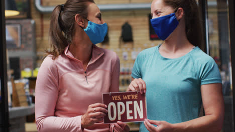 Portrait-of-caucasian-female-shopkeepers-in-face-masks-with-open-sign-in-window-of-sports-shop
