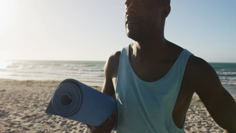 Smiling-african-american-man-holding-yoga-mat,-standing-on-beach-by-the-sea