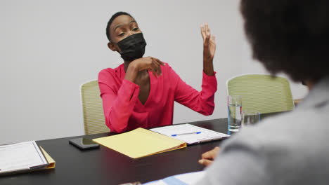 African-american-businesswoman-in-face-mask-pointing-to-screen-and-talking-to-colleagues-at-meeting