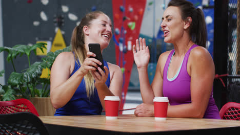 Two-happy-caucasian-women-laughing-and-looking-at-smartphone-in-cafe-at-indoor-climbing-wall