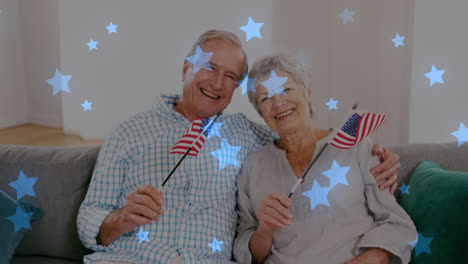 Animación-De-Estrellas-Azules-Brillantes-Sobre-El-Retrato-De-Una-Feliz-Pareja-De-Ancianos-Sosteniendo-Banderas-Americanas