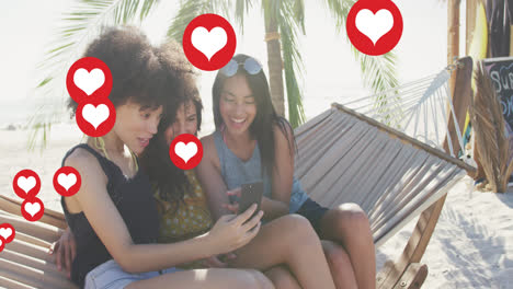 Animation-of-red-heart-love-digital-icons-over-female-friends-in-hammock-with-smartphone-on-beach